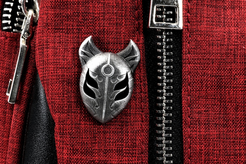 Op de afbeelding: Een zilveren metalen pin in de vorm van een vossenmasker met een zwart-wit ontwerp. De pin is bevestigd aan een rode stoffen achtergrond.