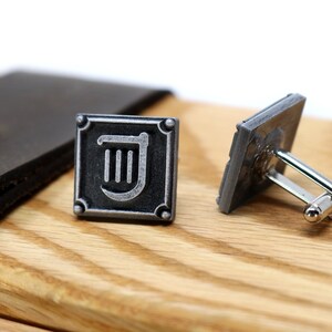 Puede incluir: Un par de gemelos de plata con un cuadrado negro y un s&iacute;mbolo de arpa plateado. Los gemelos est&aacute;n sobre una superficie de madera.