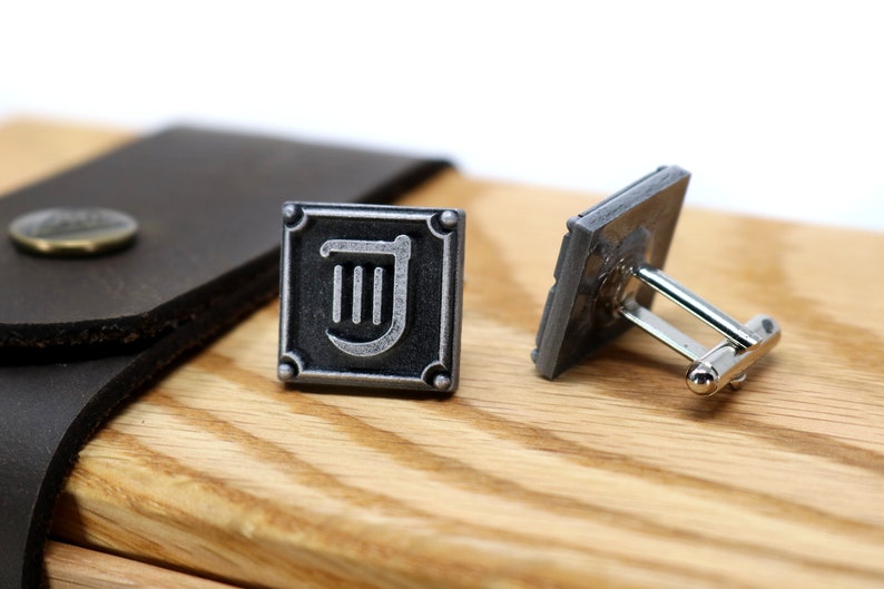 Puede incluir: Un par de gemelos de plata con un cuadrado negro y un s&iacute;mbolo de arpa plateado. Los gemelos est&aacute;n sobre una superficie de madera.