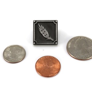 May include: A silver pin with a black square background and a silver wheat stalk design. The pin is shown next to three US coins for size comparison.