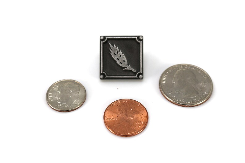 May include: A silver pin with a black square background and a silver wheat stalk design. The pin is shown next to three US coins for size comparison.