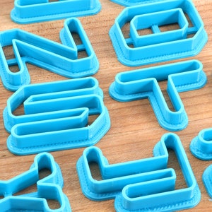 May include: A set of blue plastic cookie cutters shaped like letters of the alphabet. The cutters are arranged on a wooden surface.