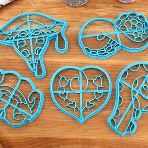 May include: Five blue plastic cookie cutters on a wooden surface. The cutters are shaped like a heart, a pizza slice, a swirl, a candy cane, and a flower.