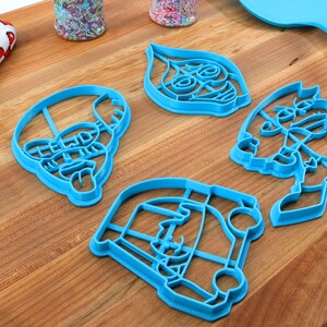 Puede incluir: Cuatro cortadores de galletas de pl&aacute;stico azul con forma de personajes de dibujos animados. Los cortadores est&aacute;n sobre una superficie de madera.
