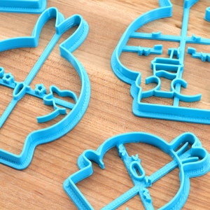 May include: Four blue plastic cookie cutters shaped like a bunny, a snowman, a penguin, and a Christmas tree. The cutters are on a wooden surface.