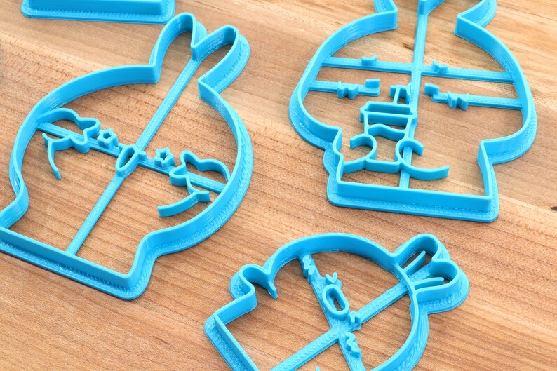 May include: Four blue plastic cookie cutters shaped like a bunny, a snowman, a penguin, and a Christmas tree. The cutters are on a wooden surface.