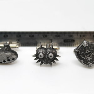 May include: A set of three silver cufflinks featuring iconic symbols from the Legend of Zelda video game series: the Ocarina of Time, Majora's Mask, and the Hylian Shield.