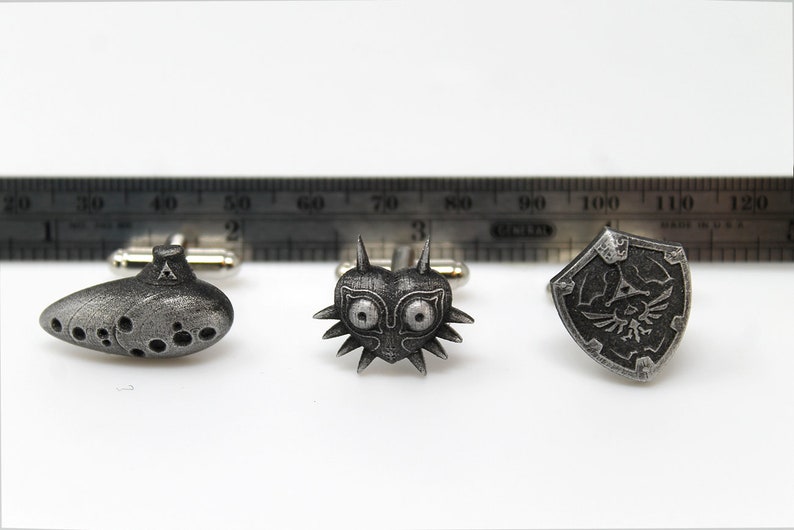 May include: A set of three silver cufflinks featuring iconic symbols from the Legend of Zelda video game series: the Ocarina of Time, Majora's Mask, and the Hylian Shield.