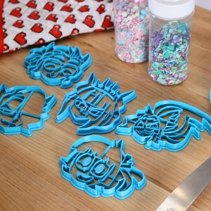May include: Five blue plastic cookie cutters shaped like cartoon characters. The cutters are on a wooden surface with sprinkles and a rolling pin in the background.