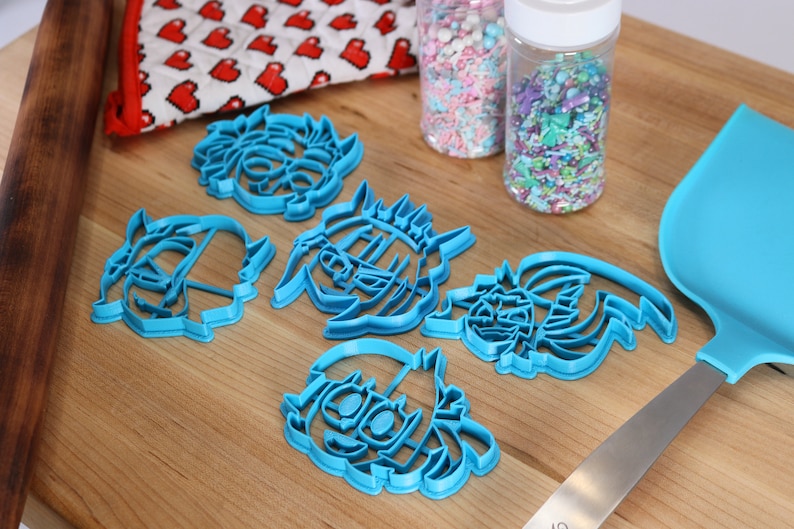 May include: Five blue plastic cookie cutters shaped like cartoon characters. The cutters are on a wooden surface with sprinkles and a rolling pin in the background.
