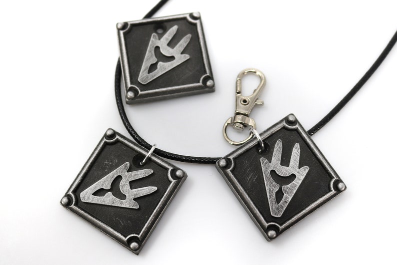 May include: Three silver-toned pendants with a black and silver design. The pendants are attached to a black cord and a silver chain. The pendants are shaped like squares with rounded corners. The design on the pendants is a stylized hand with three fingers.