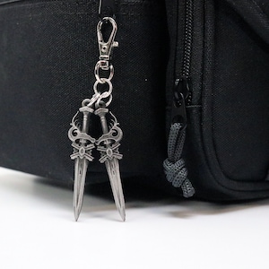 May include: A silver-colored keychain featuring two crossed swords with ornate handles and decorative elements. The keychain is attached to a black bag with a silver clasp.