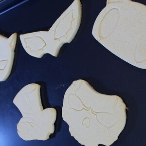 May include: Five unbaked sugar cookies on a black baking sheet. The cookies are shaped like a bat, a skull, a cat, a top hat, and a heart.