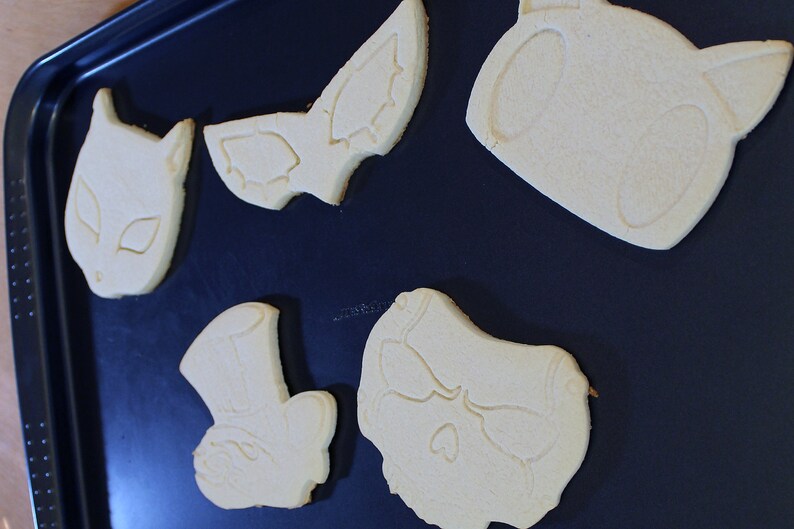 May include: Five unbaked sugar cookies on a black baking sheet. The cookies are shaped like a bat, a skull, a cat, a top hat, and a heart.