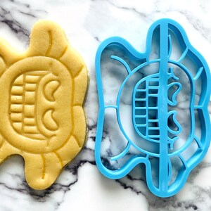 May include: A blue plastic cookie cutter and a yellow cookie in the shape of a smiling face with a wide open mouth and two eyes.