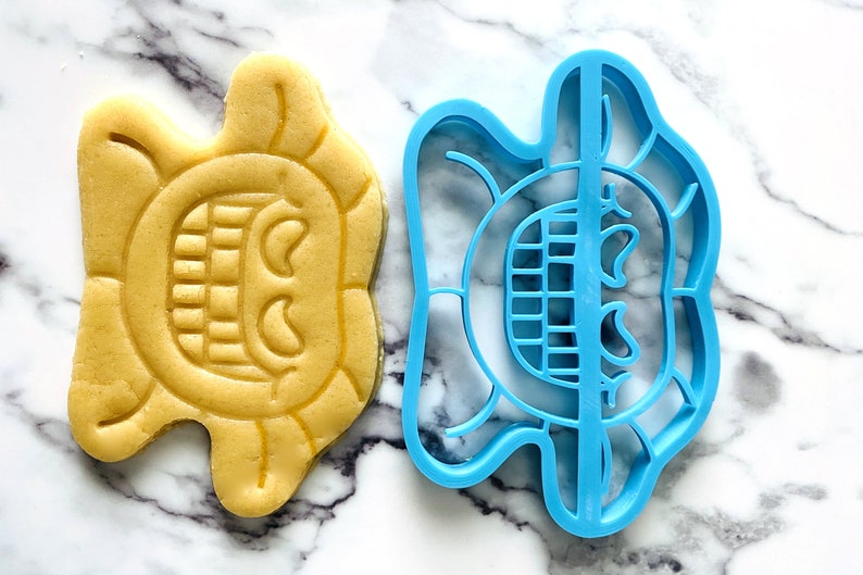 May include: A blue plastic cookie cutter and a yellow cookie in the shape of a smiling face with a wide open mouth and two eyes.
