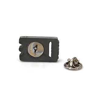 May include: A black metal pin with a silver pin back. The pin features a detailed design of a razor blade.