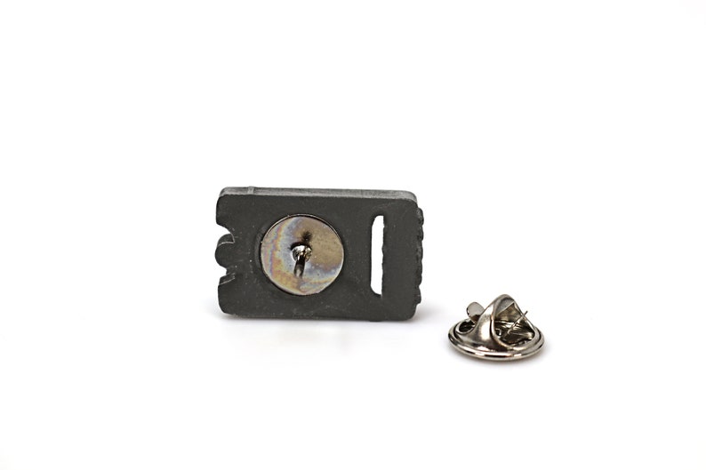 May include: A black metal pin with a silver pin back. The pin features a detailed design of a razor blade.