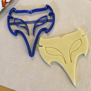 May include: A blue plastic cookie cutter in the shape of a stylized fox head next to a cutout cookie. The cookie cutter has a detailed design with leaf-like shapes. The cookie is light yellow and sits on parchment paper.