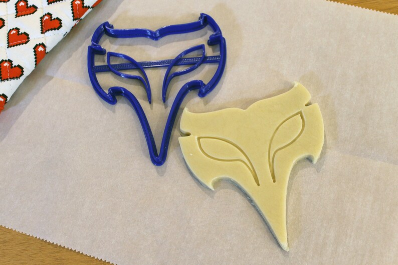 May include: A blue plastic cookie cutter in the shape of a stylized fox head next to a cutout cookie. The cookie cutter has a detailed design with leaf-like shapes. The cookie is light yellow and sits on parchment paper.