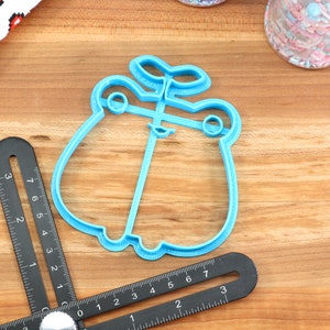 May include: A blue plastic cookie cutter shaped like a frog with a plant growing on its head. The cutter is on a wooden surface.
