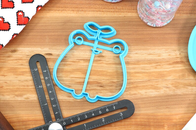 May include: A blue plastic cookie cutter shaped like a frog with a plant growing on its head. The cutter is on a wooden surface.