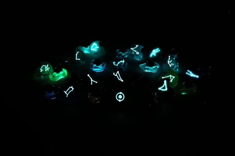 May include: A collection of glowing dice with constellations printed on each face. The dice are a variety of colors, including green, blue, and purple. The constellations are visible in the dark.