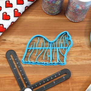 May include: A blue plastic cookie cutter shaped like a Shih Tzu dog. The cutter is on a wooden surface with a ruler and a spatula.