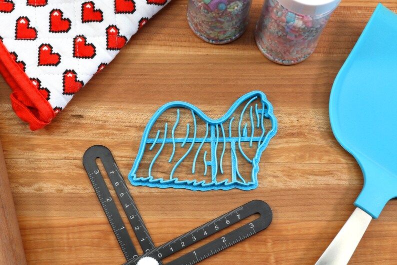 May include: A blue plastic cookie cutter shaped like a Shih Tzu dog. The cutter is on a wooden surface with a ruler and a spatula.