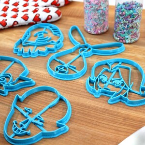 Pu&ograve; includere: Un set di cinque tagliabiscotti in plastica blu a forma di personaggi dei cartoni animati. I tagliabiscotti sono su una superficie di legno con due barattoli di codette colorate sullo sfondo.