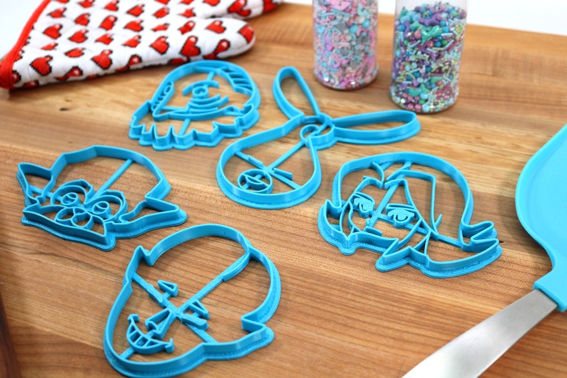 Pu&ograve; includere: Un set di cinque tagliabiscotti in plastica blu a forma di personaggi dei cartoni animati. I tagliabiscotti sono su una superficie di legno con due barattoli di codette colorate sullo sfondo.