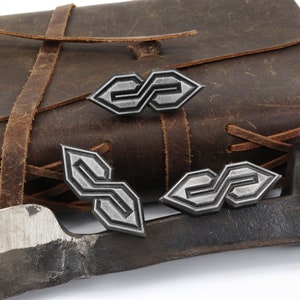 May include: A collection of silver-colored metal pins with an interlocking "S" design, displayed near a brown leather satchel and a piece of metal. The pins have a geometric pattern and are likely decorative.