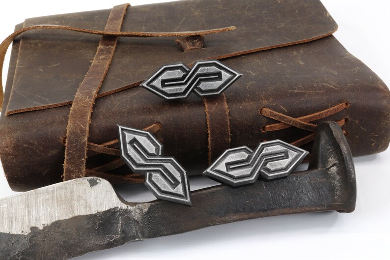 May include: A collection of silver-colored metal pins with an interlocking "S" design, displayed near a brown leather satchel and a piece of metal. The pins have a geometric pattern and are likely decorative.