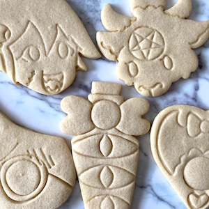 Pode incluir: Cinco biscoitos simples de a&ccedil;&uacute;car decorados com v&aacute;rios desenhos. Os desenhos incluem um rosto de desenho animado, um cora&ccedil;&atilde;o com um la&ccedil;o, uma garrafa com um la&ccedil;o, um morcego com um la&ccedil;o e um dem&ocirc;nio com um pentagrama.