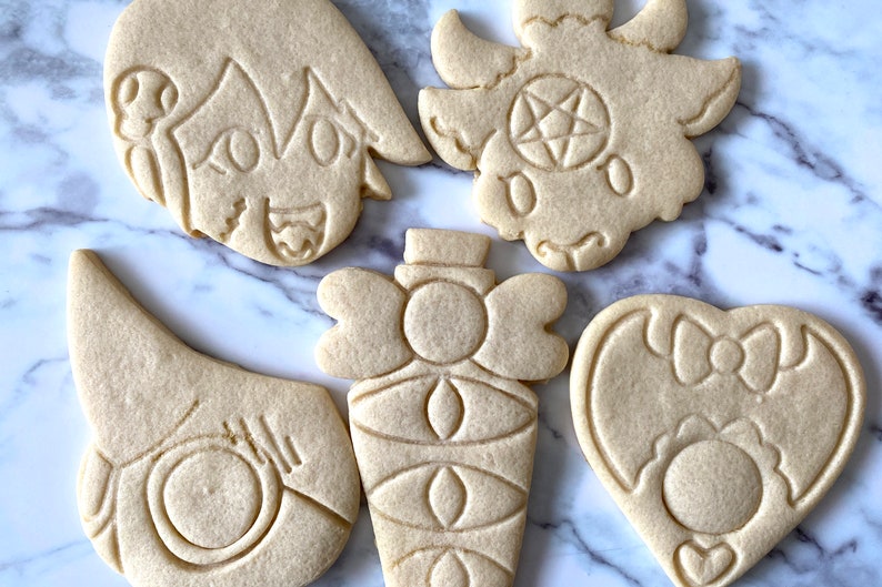 Pode incluir: Cinco biscoitos simples de a&ccedil;&uacute;car decorados com v&aacute;rios desenhos. Os desenhos incluem um rosto de desenho animado, um cora&ccedil;&atilde;o com um la&ccedil;o, uma garrafa com um la&ccedil;o, um morcego com um la&ccedil;o e um dem&ocirc;nio com um pentagrama.