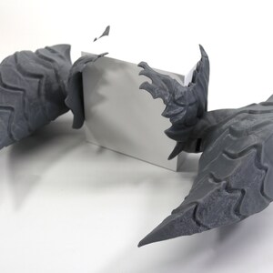 Op de afbeelding: Een 3D-geprint model van een wezen met twee grote, grijze vleugels. De vleugels zijn getextureerd en hebben een scherpe, puntige vorm. Het model staat op een wit oppervlak.
