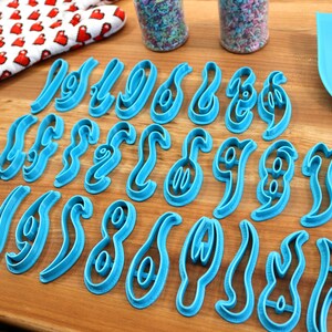 May include: A set of 26 blue plastic cookie cutters shaped like the numbers 0-9 in a cursive font. The cutters are arranged on a wooden surface.