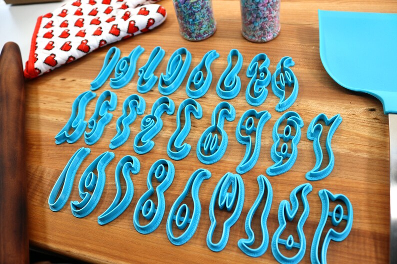 May include: A set of 26 blue plastic cookie cutters shaped like the numbers 0-9 in a cursive font. The cutters are arranged on a wooden surface.