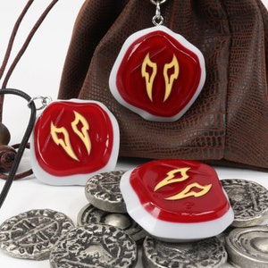 May include: Three red and white resin pendants with a gold design, hanging from a black cord and a brown leather pouch. The pendants are shaped like a shield with a white border and a red centre. The gold design is a stylised flame shape. The pendants are surrounded by silver coins.