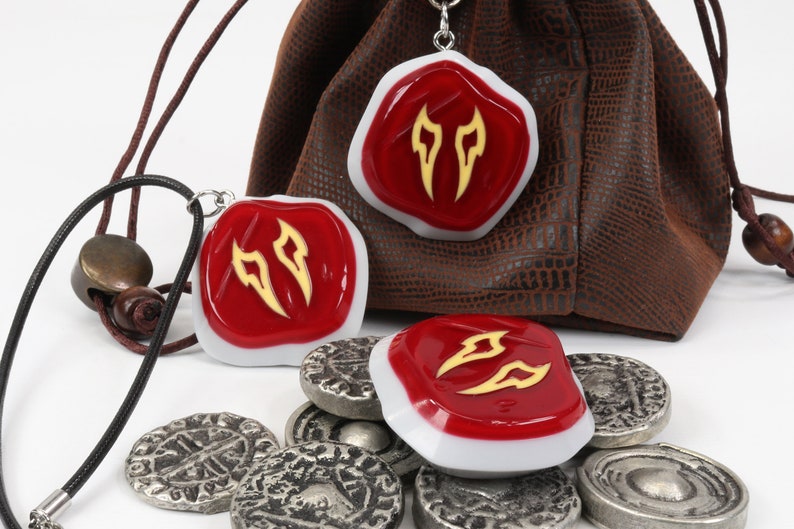 May include: Three red and white resin pendants with a gold design, hanging from a black cord and a brown leather pouch. The pendants are shaped like a shield with a white border and a red centre. The gold design is a stylised flame shape. The pendants are surrounded by silver coins.