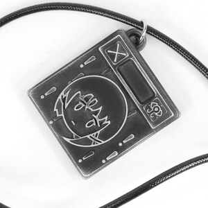 May include: A black and silver metal pendant necklace with a square charm featuring a cartoon face and the symbols 'X' and '!' on the charm.