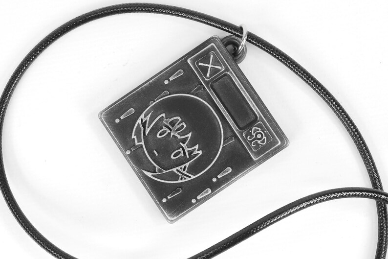 May include: A black and silver metal pendant necklace with a square charm featuring a cartoon face and the symbols 'X' and '!' on the charm.