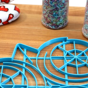 May include: A blue plastic circular cookie cutter with a web-like design. The cutter is on a wooden surface with two jars of colorful sprinkles in the background.
