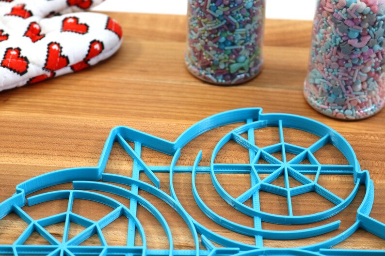 May include: A blue plastic circular cookie cutter with a web-like design. The cutter is on a wooden surface with two jars of colorful sprinkles in the background.