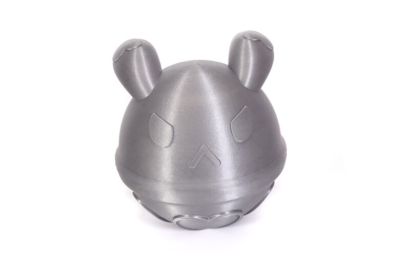 Op de afbeelding: Een zilverkleurig, 3D-geprint speelgoed met een afgeronde vorm en twee kleine oren. Het speelgoed heeft een gezicht met twee ogen en een kleine mond.