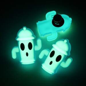 May include: Three glow-in-the-dark figurines of a character with a round head, two eyes, and a mouth. The character is wearing a hat and has two arms. The figurines are made of a soft, flexible material and are a light green colour.