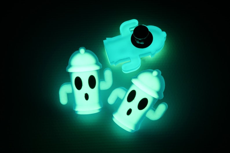 May include: Three glow-in-the-dark figurines of a character with a round head, two eyes, and a mouth. The character is wearing a hat and has two arms. The figurines are made of a soft, flexible material and are a light green colour.