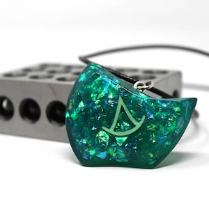 Puede incluir: Un colgante de resina iridiscente verde y azul con un s&iacute;mbolo blanco, en un collar de cord&oacute;n negro.