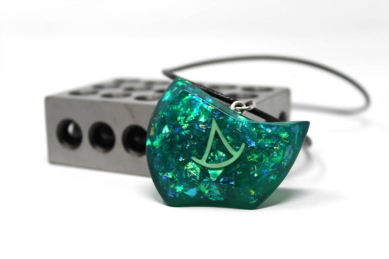 Puede incluir: Un colgante de resina iridiscente verde y azul con un s&iacute;mbolo blanco, en un collar de cord&oacute;n negro.