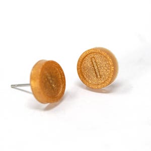 May include: A pair of gold-colored stud earrings with a textured surface. Each earring features a raised, horizontal line.